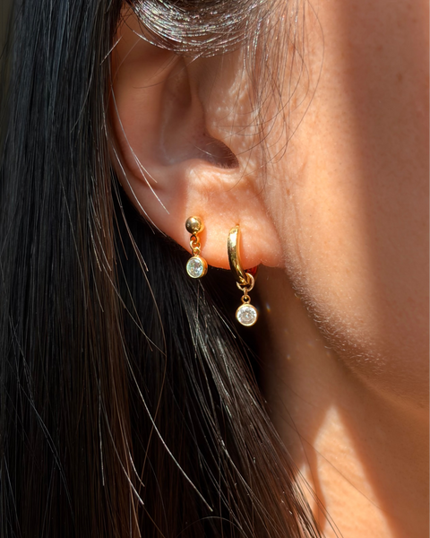 A close-up of a person's ear wearing two small gold hoop earrings with round clear stones, shown in bright natural light against dark hair.