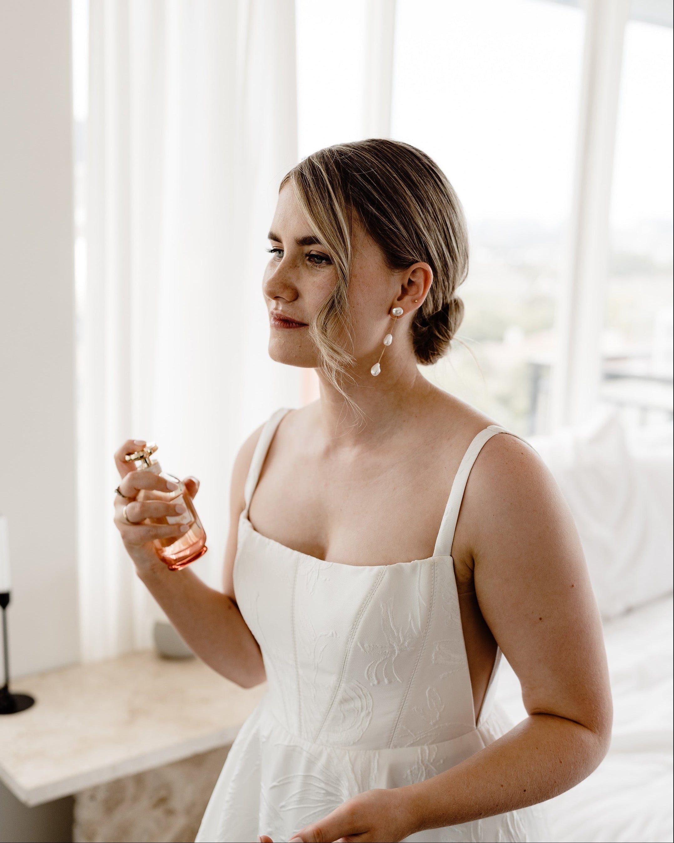 Woman in a white dress holding a perfume bottle in a softly lit room.