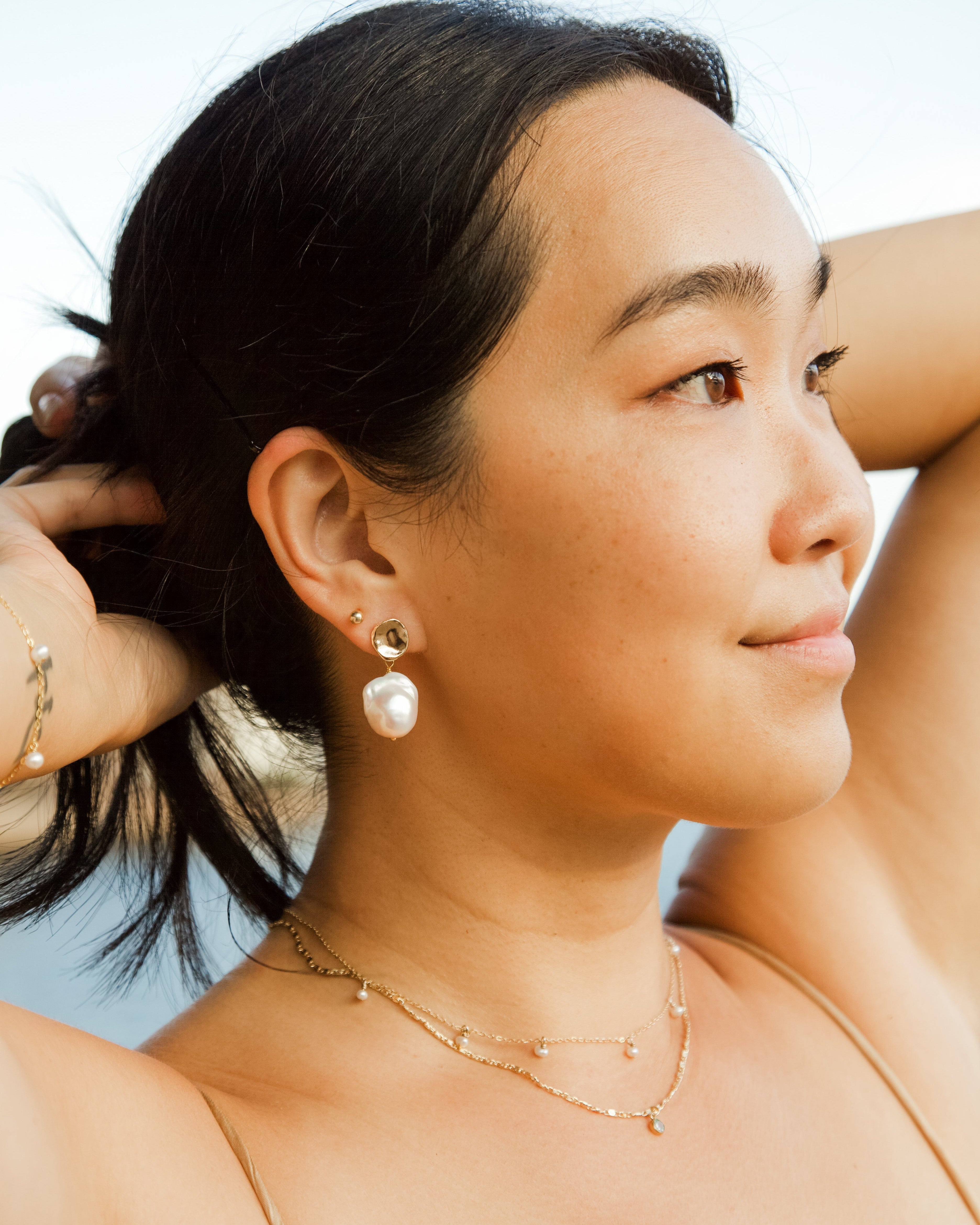 Woman with hair tied back, wearing pearl earrings and a pearl necklace, against a clear sky.