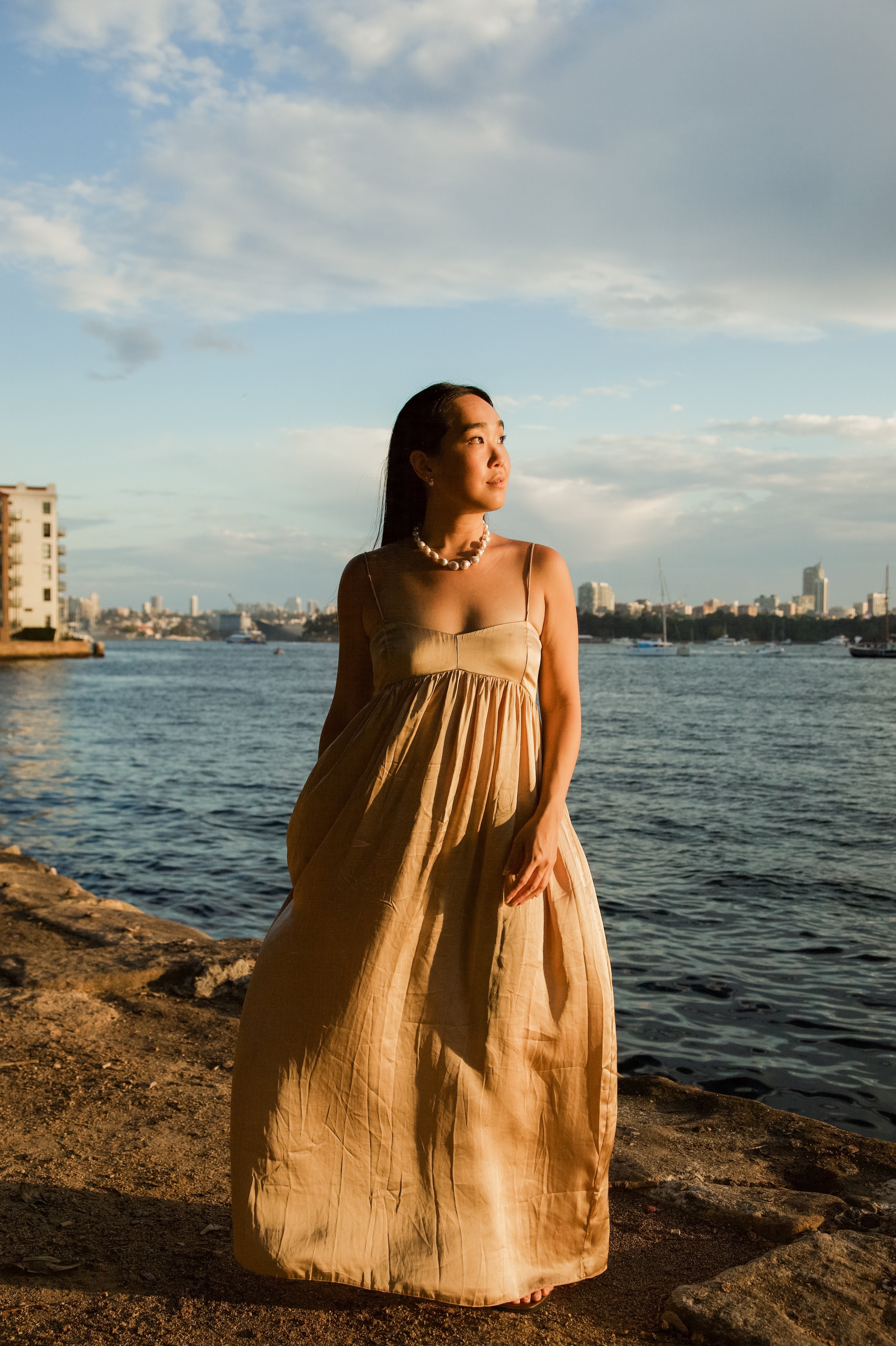 Woman in a beige dress standing by a waterfront with a city skyline in the background.
