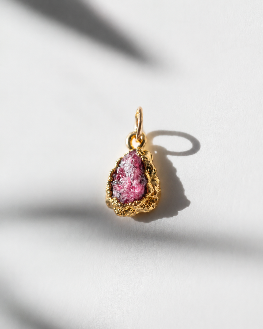 A gold teardrop pendant featuring a raw, textured ruby, photographed on a white surface with dramatic leaf shadows.
