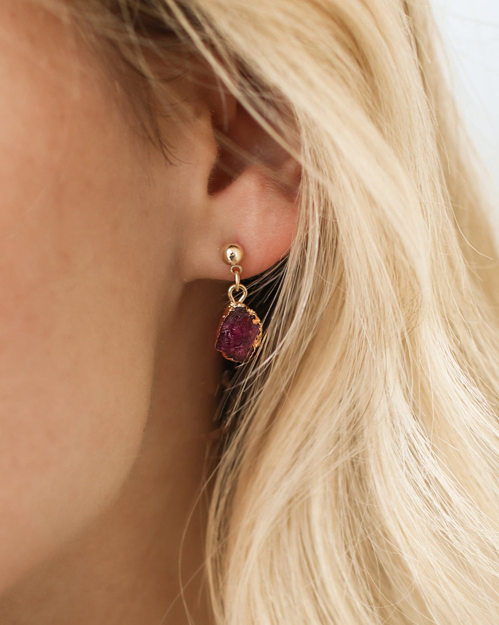 A person with blonde hair wearing gold stud earrings with red gemstones, potentially the July birthstone ruby.